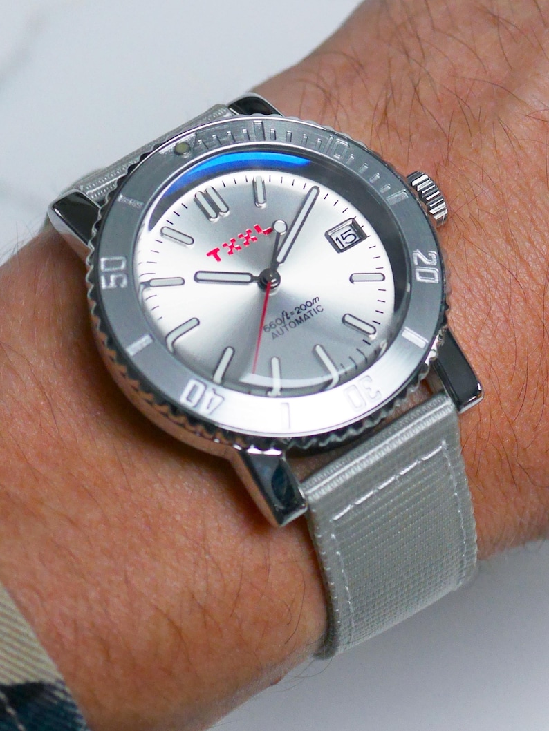 Puede incluir: Un reloj de pulsera plateado con una correa de nailon gris. La esfera del reloj es plateada con marcas negras y una aguja de segundos roja. La esfera del reloj tambi&eacute;n tiene una ventana de fecha y el texto "160ft/200m AUTOMATIC" en rojo.