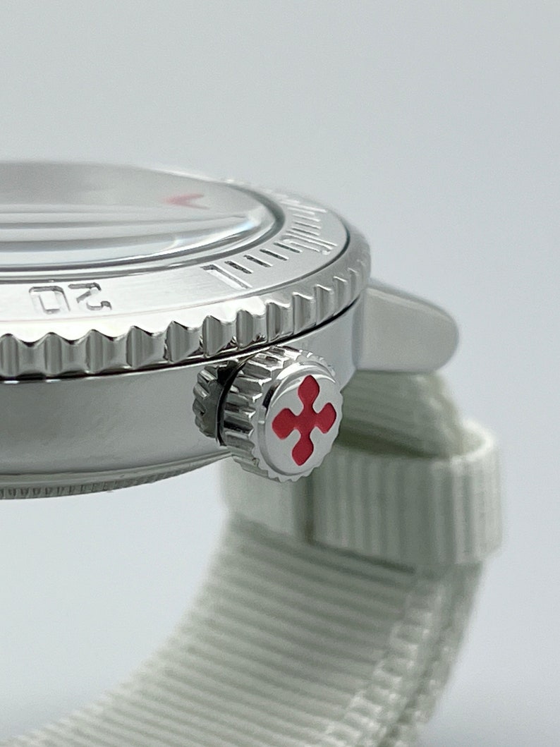 Puede incluir: Un reloj de pulsera plateado con una banda blanca y una cruz roja en la corona.