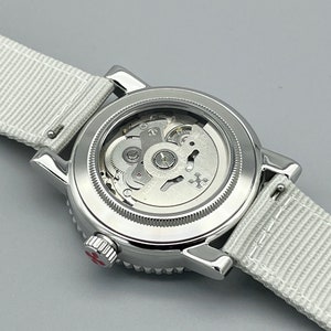 Puede incluir: Un reloj de pulsera plateado con una banda de tela blanca. La esfera del reloj es visible y muestra el funcionamiento interno del reloj.