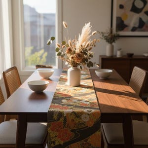 May include: A dining table set with a floral patterned table runner, two white bowls, and a vase of dried flowers. The table is made of dark wood and has four chairs with woven backs and gray seats. A framed abstract art piece hangs on the wall.