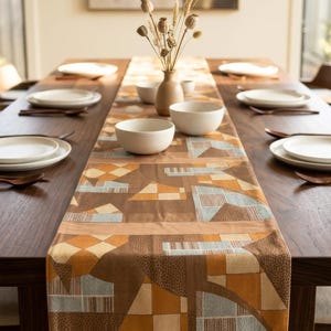 Puede incluir: Un camino de mesa rectangular largo con un patrón geométrico en tonos marrón, beige y azul claro. La tela muestra una mezcla de cuadrados sólidos, cuadrículas texturizadas y formas triangulares. Está centrado sobre una mesa de madera oscura.