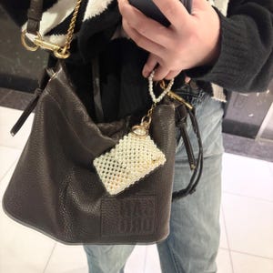 以下が含まれることがあります： ゴールドチェーンストラップと小さなパールビーズの携帯電話ケースが付いた、濃い茶色のレザーバケットバッグ。携帯電話ケースは白くて長方形です。バッグは質感のある表面と巾着開閉式です。