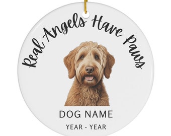 May include: White ceramic ornament with a golden hanging cord. The ornament features a brown dog illustration and the text "Real Angels Have Paws". Below the dog is the text "DOG NAME" and "YEAR - YEAR".