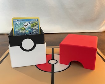 Puede incluir: Un tarjetero blanco y negro con un diseño circular, que contiene una tarjeta coleccionable con un personaje de dibujos animados. Un tarjetero rojo está al lado, sobre un tapete de juego marrón y rojo. La tarjeta dice "Oshawott".