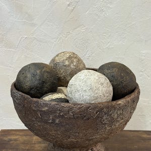 May include: A dark brown, textured pedestal bowl filled with several spherical objects. The spheres vary in colour, including solid dark charcoal, light grey, and off-white, with some featuring visible paper-like text fragments on their surfaces.