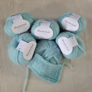 Könnte beinhalten: Fünf Knäuel hellblaues Wonderfluff Bulky Weight Garn, jeweils mit einem weißen Papieretikett, angeordnet um ein kleines, rechteckiges Strickmuster aus demselben hellblauen Garn mit einer strukturierten, flauschigen Oberfläche.