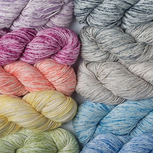 May include: A selection of colourful yarn skeins in shades of purple, pink, orange, yellow, green, blue, and grey. The yarn has a soft, textured appearance, suitable for knitting or crochet. The skeins are arranged closely together, showcasing the variety of colours.