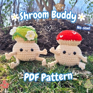 May include: Two crocheted mushroom figures with smiling faces and outstretched arms. One has a green cap with white flowers, the other a red cap with white hearts. Text reads "Shroom Buddy" and "PDF Pattern".