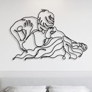 Modern metal wall decor illustrating an intimate couple embrace in black line art, hanging above a contemporary minimalist bed with white linens.