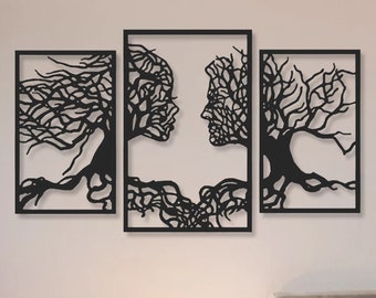 Op de afbeelding: Een zwarte metalen wandkunst sculptuur met drie delen die gestileerde bomen voorstellen met in elkaar verweven takken die de vorm van twee gezichten vormen. De sculptuur is gemonteerd op een witte muur boven een bed met witte beddengoed.