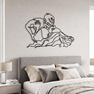 Minimalist metal wall art of a kissing couple in a matte black finish, centered perfectly above a grey upholstered bed in a bright modern bedroom.