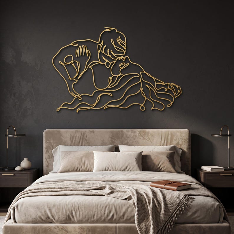 Luxury metal wall art depicting a minimalist couple kissing in a premium gold finish, mounted above a plush velvet bed in a moody modern bedroom.