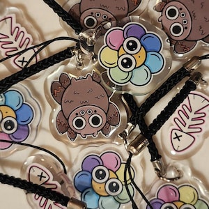 May include: A set of colorful keychains featuring various designs, including a spider, a flower, and a fish bone. The keychains are made of clear plastic with colorful graphics and are attached to black cords.