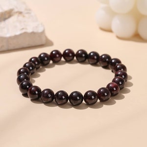 May include: A dark red beaded bracelet made of round, polished stones. The bracelet is made of natural gemstones and is a popular choice for jewellery.