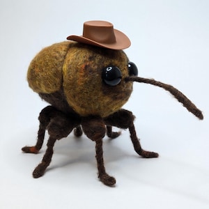 Peut inclure: Un scarabée en feutre brun et orange portant un chapeau de cowboy brun avec des yeux noirs et de longues pattes brunes.