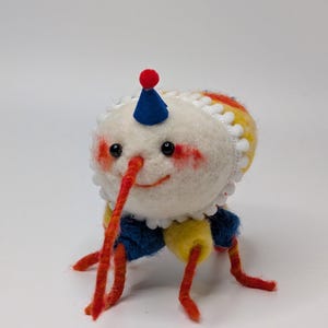 May include: A whimsical felt creature with a white head, adorned with a blue party hat and red accents. It has a long, orange nose and matching legs. The creature's body features blue and yellow sections, creating a playful, handmade aesthetic.