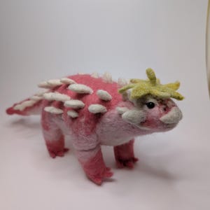 Może przedstawiać: Ręcznie wykonana różowo-biała filcowa figurka dinozaura z białymi kolcami wzdłuż grzbietu i zielonym pękiem na głowie. Dinozaur ma małe, ciemne oczy i stoi na czterech nogach.