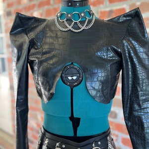 May include: A black faux leather cropped jacket with a crocodile print and exaggerated shoulders. The jacket has a V-shaped neckline and is worn with a silver chain choker and a black leather belt with silver chain accents.