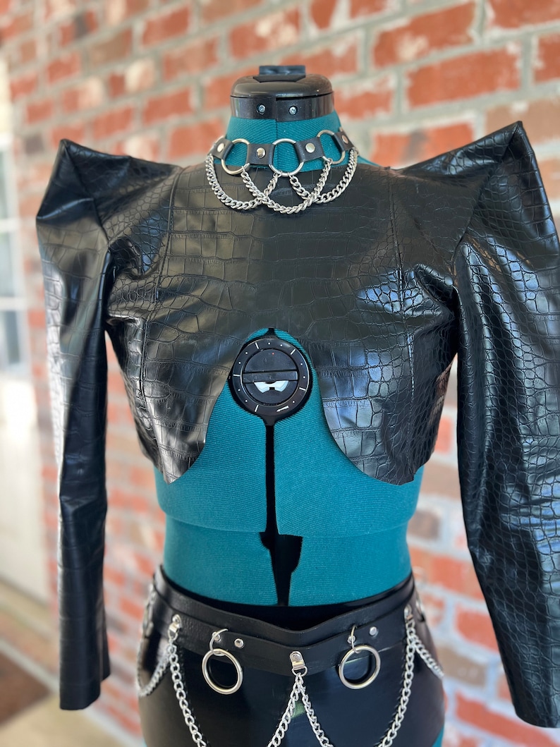 May include: A black faux leather cropped jacket with a crocodile print and exaggerated shoulders. The jacket has a V-shaped neckline and is worn with a silver chain choker and a black leather belt with silver chain accents.