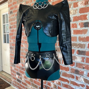 May include: A black faux leather cropped jacket with a crocodile pattern and exaggerated shoulders. The jacket has a silver chain choker and a matching silver chain belt with large rings.
