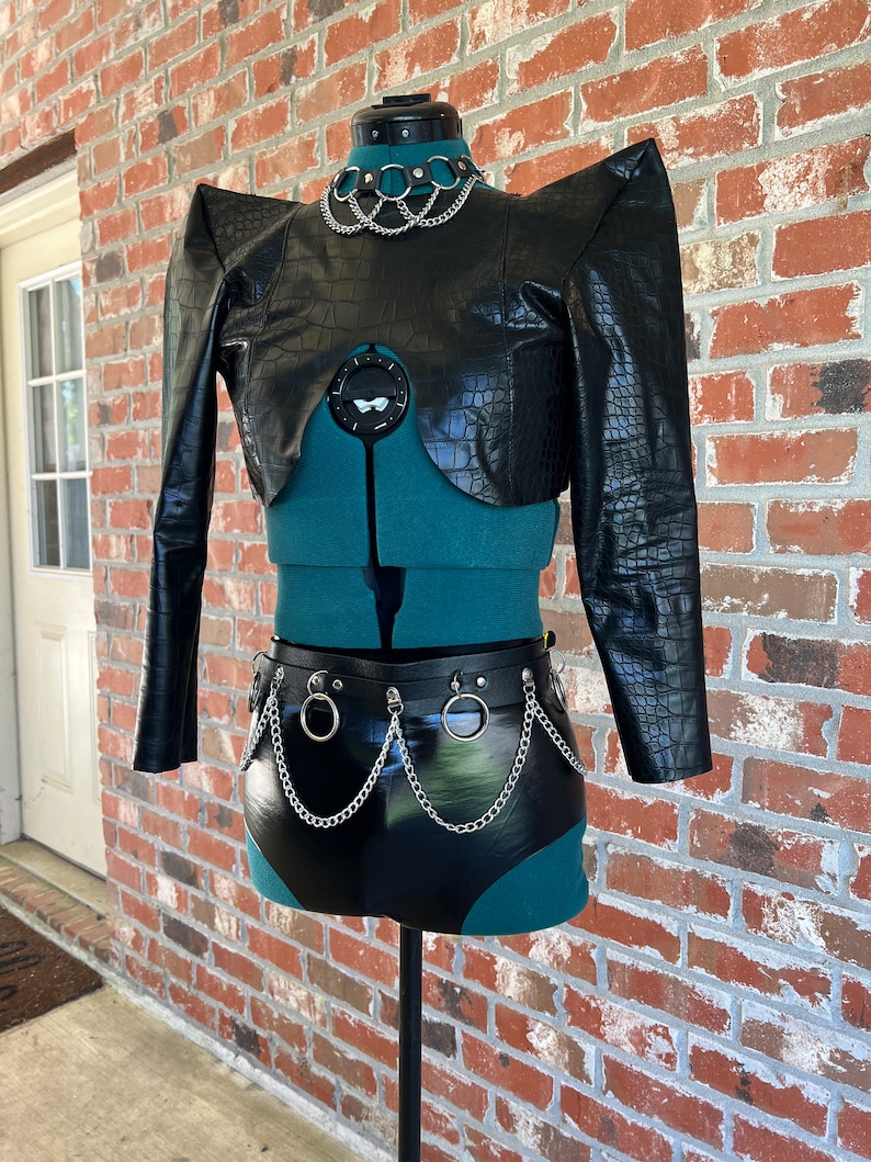 May include: A black faux leather cropped jacket with a crocodile pattern and exaggerated shoulders. The jacket has a silver chain choker and a matching silver chain belt with large rings.