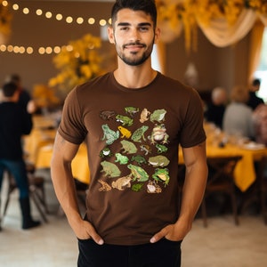May include: A brown t-shirt with a pattern of various green, yellow, and brown frogs. The shirt has a crew neck and short sleeves.