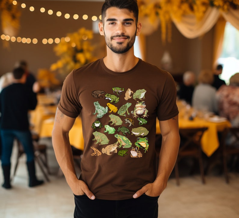 May include: A brown t-shirt with a pattern of various green, yellow, and brown frogs. The shirt has a crew neck and short sleeves.