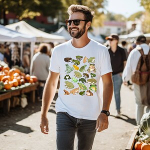 May include: A white t-shirt with a colorful pattern of frogs. The shirt features a variety of frog designs, including green, brown, and yellow frogs.