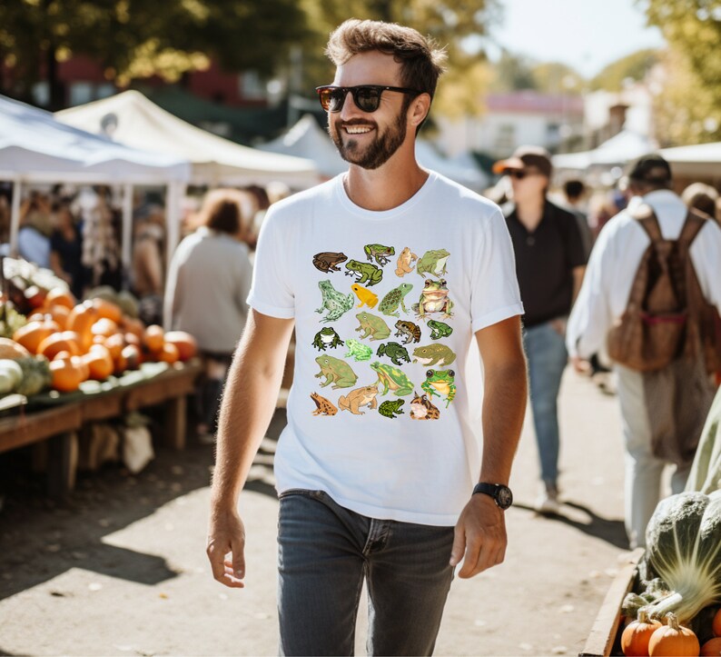 May include: A white t-shirt with a colorful pattern of frogs. The shirt features a variety of frog designs, including green, brown, and yellow frogs.