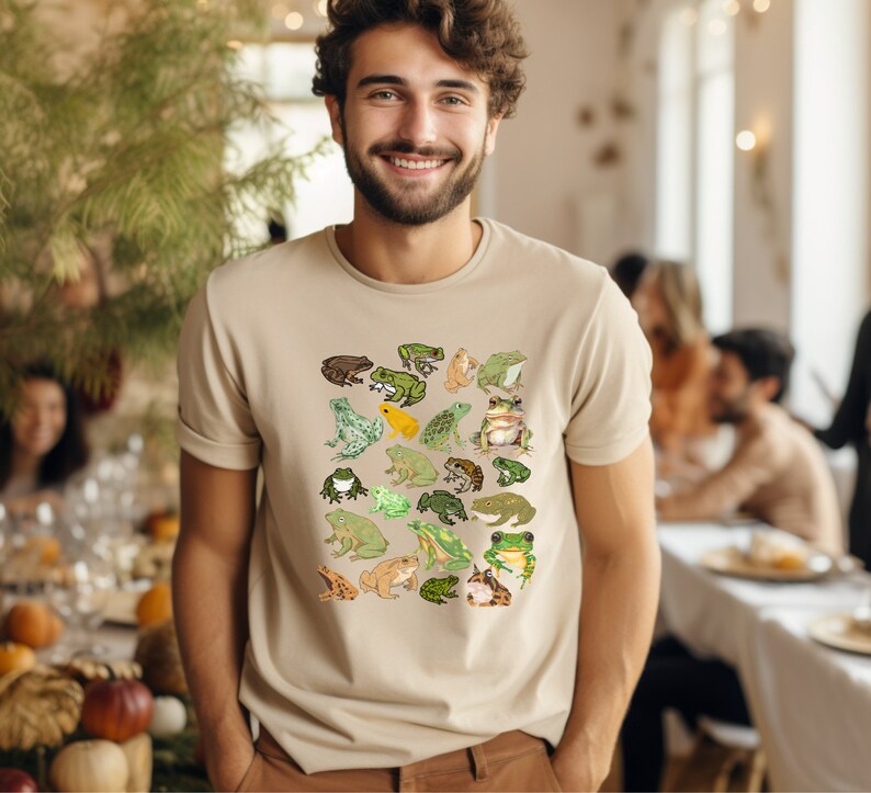 May include: A beige t-shirt with a pattern of green, brown, and yellow frogs. The frogs are arranged in a grid pattern on the front of the shirt.