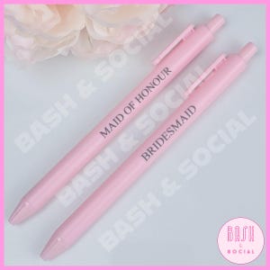 May include: Two pale pink pens with the words "MAID OF HONOUR" and "BRIDESMAID" printed in black. The pens are on a white surface with a pink border. A pink flower is in the background. The pens are from Bash & Social.