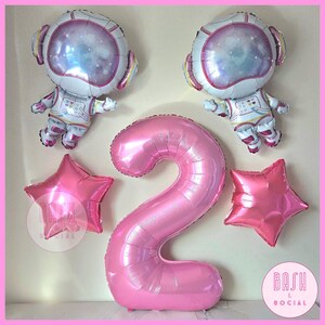 May include: A party balloon set with two astronaut-shaped balloons, a large pink number 2 balloon, and two pink star-shaped balloons. The astronaut balloons have a space theme. Perfect for a birthday party.