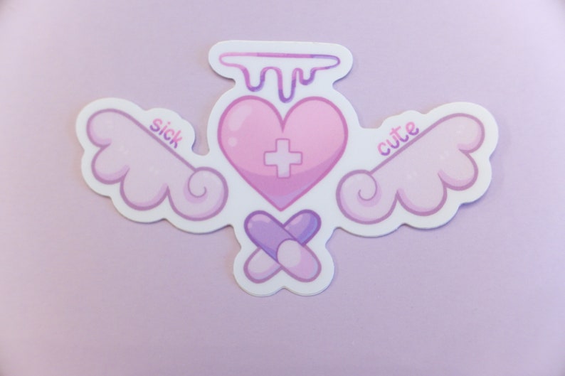 May include: A sticker featuring a pink heart with a cross, wings, and the words "sick" and "cute". Two purple pills are crossed below. The design has a white border and is set against a pale purple backdrop.