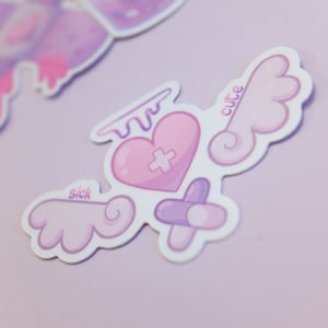 May include: A sticker featuring a pink heart with a cross, angel wings, and a halo. The sticker has the words "sick" and "cute" on it. The colour palette includes light pink, purple, and white. The design is whimsical and cute.