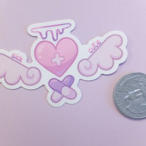 May include: A sticker featuring a pink heart with a cross, wings, and pills. The heart has a dripping design at the top. The words "sick" and "cute" are on the wings. A coin is next to the sticker.