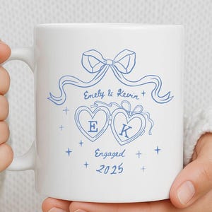 May include: White ceramic mug with a blue design featuring a bow, ribbon, and heart-shaped lockets with the letters "E" and "K." The mug also includes the text "Emely & Kevin," "Engaged," and the year "2025."