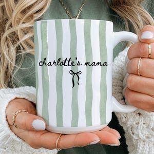 May include: White ceramic mug with vertical green and white stripes. The mug has the text "charlotte's mama" in black script, with a black bow graphic underneath. The mug is held by a person wearing a white sweater.