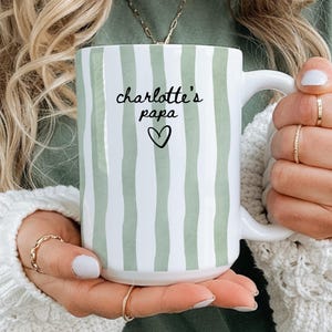 May include: White ceramic mug with vertical light green stripes. The mug features the text "charlotte's papa" in black script, with a heart symbol below. The mug has a white handle and is held by a person.