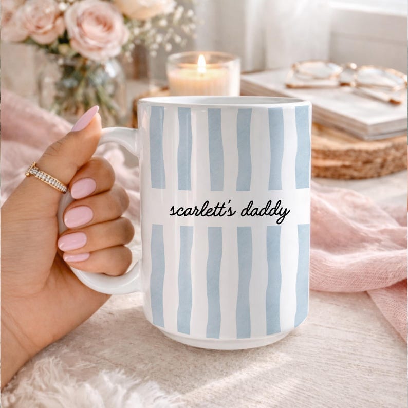 Puede incluir: Taza de cer&aacute;mica blanca con rayas verticales de color azul claro. La taza tiene el texto "scarlett's daddy" en una fuente negra. La taza est&aacute; sostenida por una mano con un anillo dorado y esmalte de u&ntilde;as rosa.