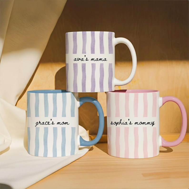 Puede incluir: Tres tazas de cer&aacute;mica con patrones de rayas verticales en colores pastel. Una taza es lavanda y blanca con "ava's mama". Otra es azul y blanca con "grace's mom" y la &uacute;ltima es rosa y blanca con "sophia's mommy".