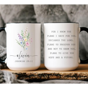 May include: White ceramic mug with a black handle and rim. The mug features a floral design with the name "Olivia" and the Bible verse Jeremiah 29:11. The opposite side has the full verse printed in black.