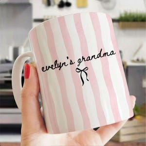 May include: A white ceramic mug with vertical pink stripes. The mug has a white handle and features the text "evelyn's grandma" in black script, with a black bow graphic. The mug is held by a person.
