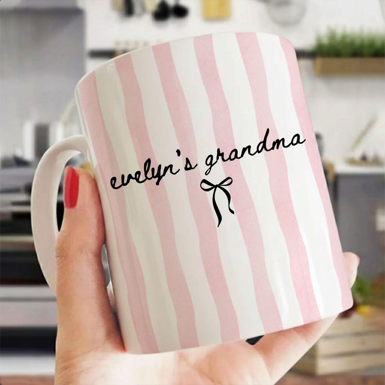 May include: A white ceramic mug with vertical pink stripes. The mug has a white handle and features the text "evelyn's grandma" in black script, with a black bow graphic. The mug is held by a person.