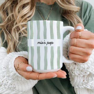 Puede incluir: Taza de cer&aacute;mica blanca con un dise&ntilde;o de rayas verdes y blancas. La taza tiene el texto "mia's pop" en una fuente manuscrita. La taza es sostenida por una persona que lleva un su&eacute;ter blanco y anillos dorados.