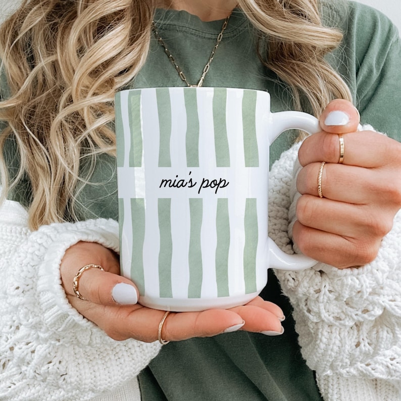 Puede incluir: Taza de cer&aacute;mica blanca con un dise&ntilde;o de rayas verdes y blancas. La taza tiene el texto "mia's pop" en una fuente manuscrita. La taza es sostenida por una persona que lleva un su&eacute;ter blanco y anillos dorados.