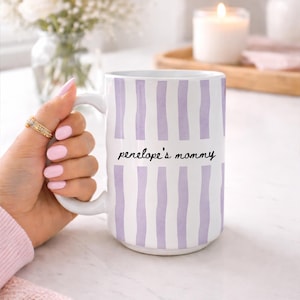 Puede incluir: Taza de cer&aacute;mica blanca con rayas verticales lavanda y el texto "penelope's mommy" en escritura negra. La taza tiene un asa grande. La taza es sostenida por una persona con esmalte de u&ntilde;as rosa y anillos dorados.