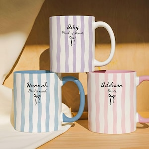 May include: Three ceramic mugs with vertical striped patterns in pastel colors: lavender, blue, and pink. Each mug has a different handle color and text: "Riley Maid of Honor", "Hannah Bridesmaid", and "Addison Bride". A black bow graphic is below each name.