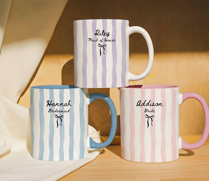May include: Three ceramic mugs with vertical striped patterns in pastel colors: lavender, blue, and pink. Each mug has a different handle color and text: "Riley Maid of Honor", "Hannah Bridesmaid", and "Addison Bride". A black bow graphic is below each name.