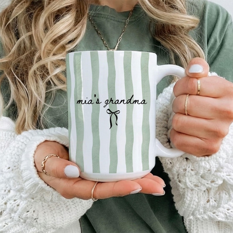 White ceramic coffee mug with soft green vertical stripes and elegant black script showing grandchild name and a small bow detail, created as a pregnancy announcement gift for a new grandma.grandparents.