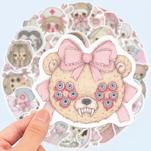 May include: A collection of stickers featuring a variety of eerie, pastel-colored illustrations. The central sticker is a beige teddy bear with multiple blue eyes, a pink bow, and sharp teeth. Other stickers include dolls and figures in pink and white.